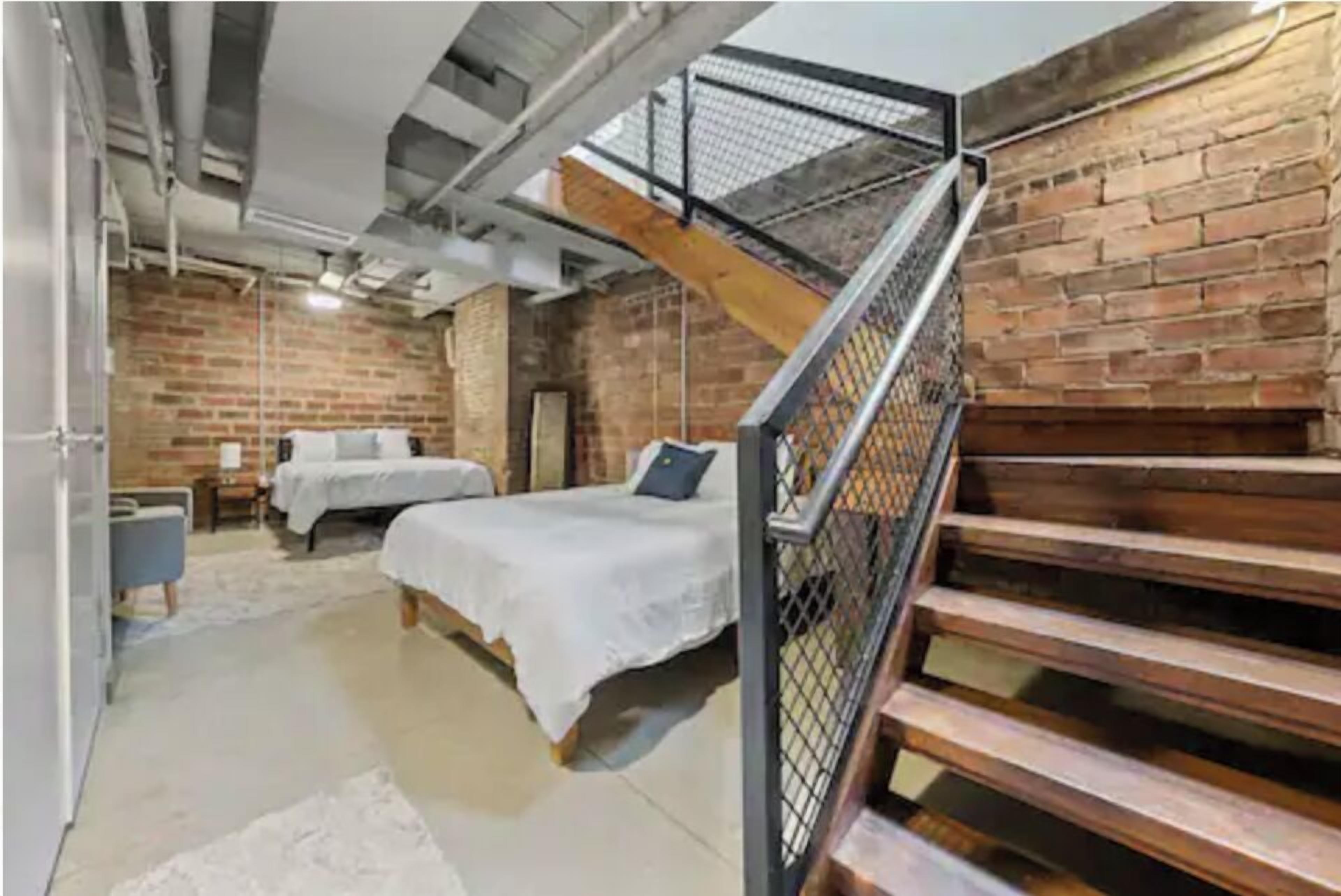 Een kamer met een bakstenen muur, een trap met een metalen leuning, twee bedden en een witte vloerkleed.