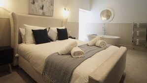 Un letto rifatto con cuscini, un comodino, una lampada a parete, uno specchio rotondo, un portasciugamani e un bagno con lavandino e vasca.