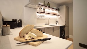 Una cucina moderna con un piano di lavoro, un tagliere con pane e formaggio, un coltello, una lavatrice e uno scaffale con bottiglie e bicchieri.