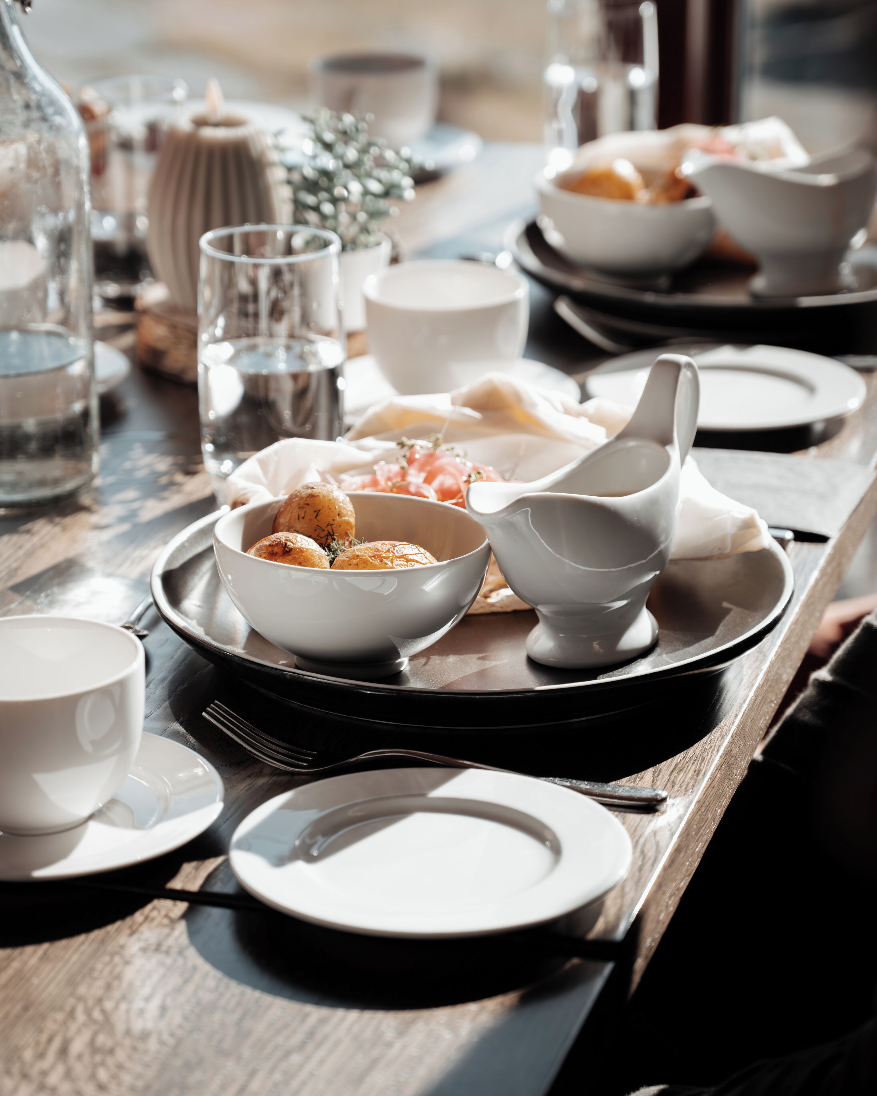 A dining table set with various dishes, including a bowl of roasted potatoes, a white bowl, and several white cups and saucers.