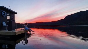 Un muelle con una silla y un flotador salvavidas, una persona sentada junto al agua, y una puesta de sol sobre un lago tranquilo con montañas al fondo.