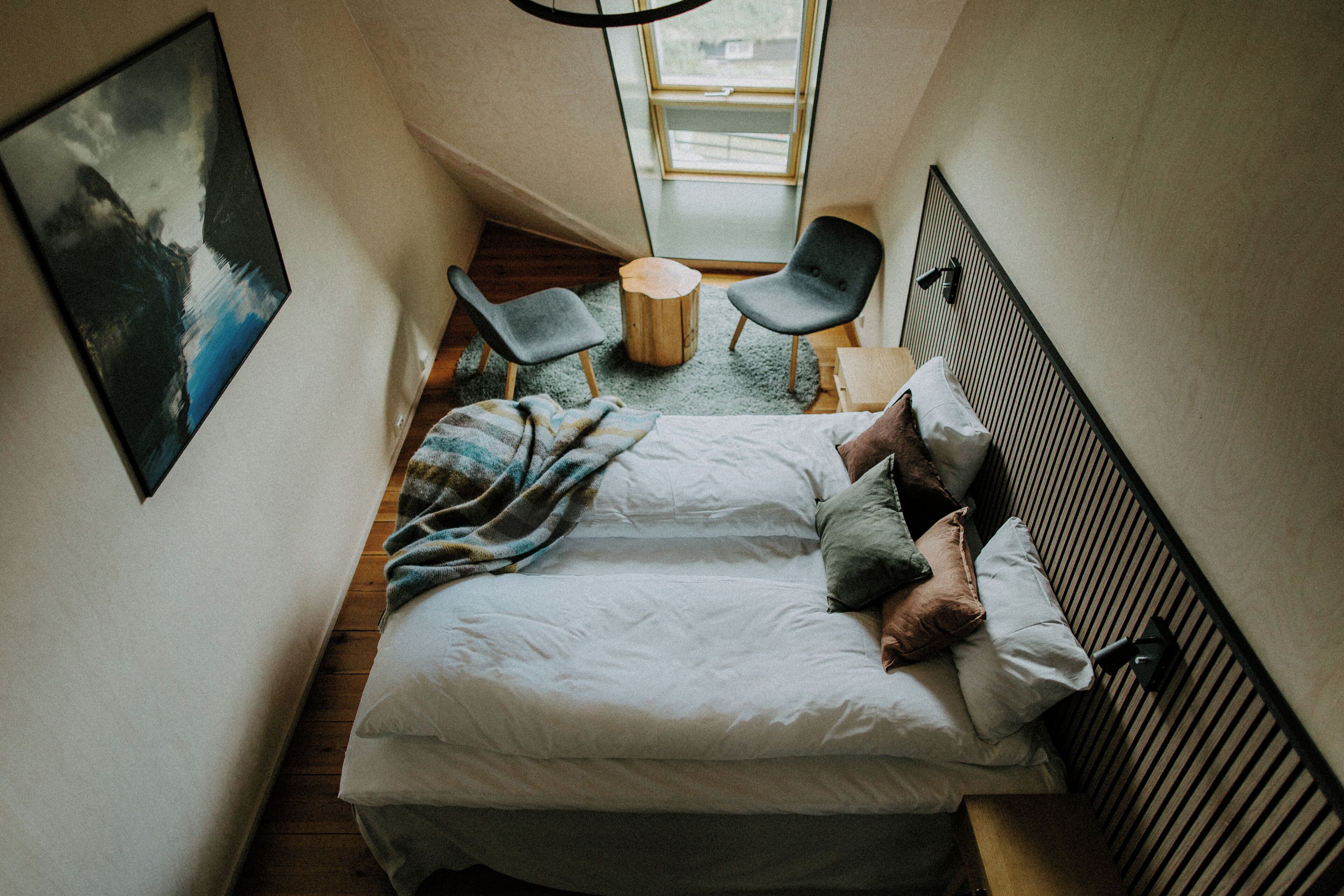 Una habitación estrecha con una cama, dos sillas, un taburete de madera y un cuadro en la pared.