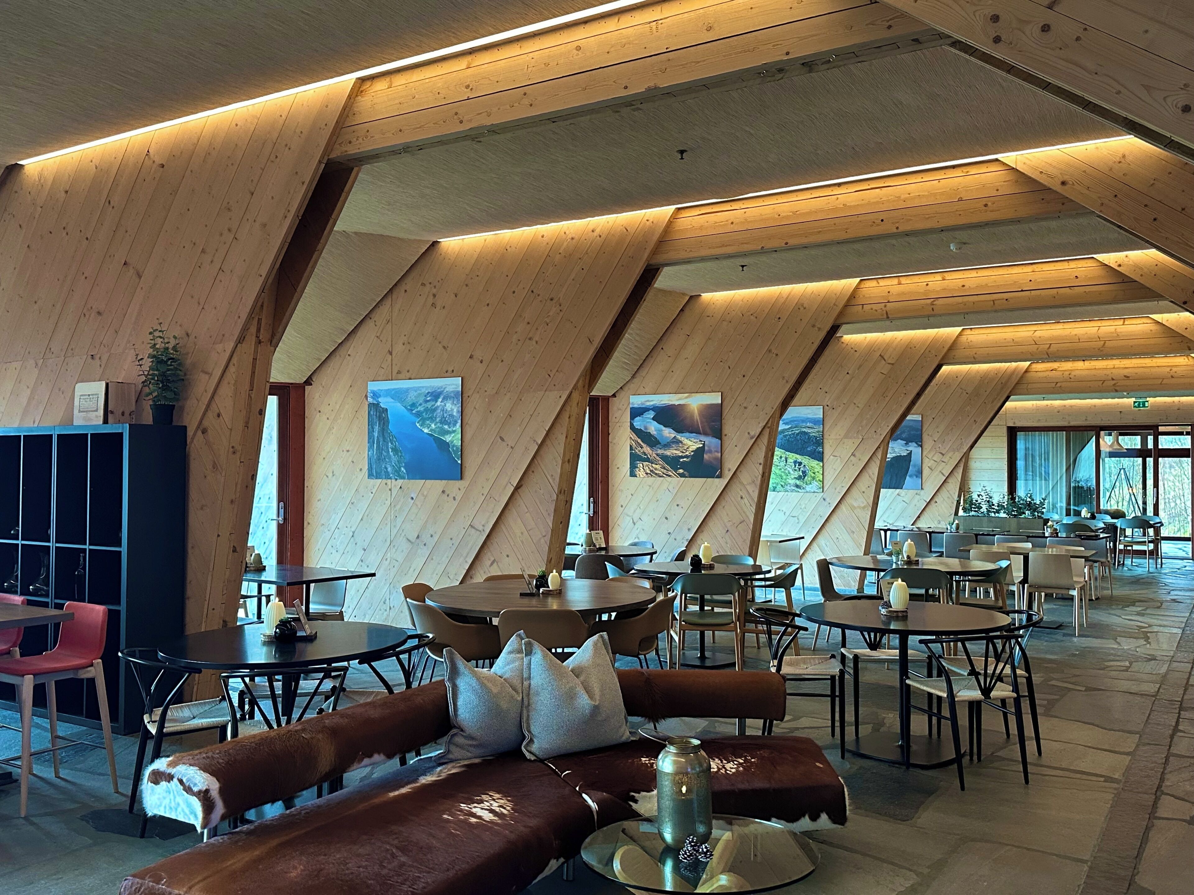 Un interior moderno con techos inclinados de madera, varias mesas y sillas de comedor, y ventanas grandes que ofrecen vistas al exterior.