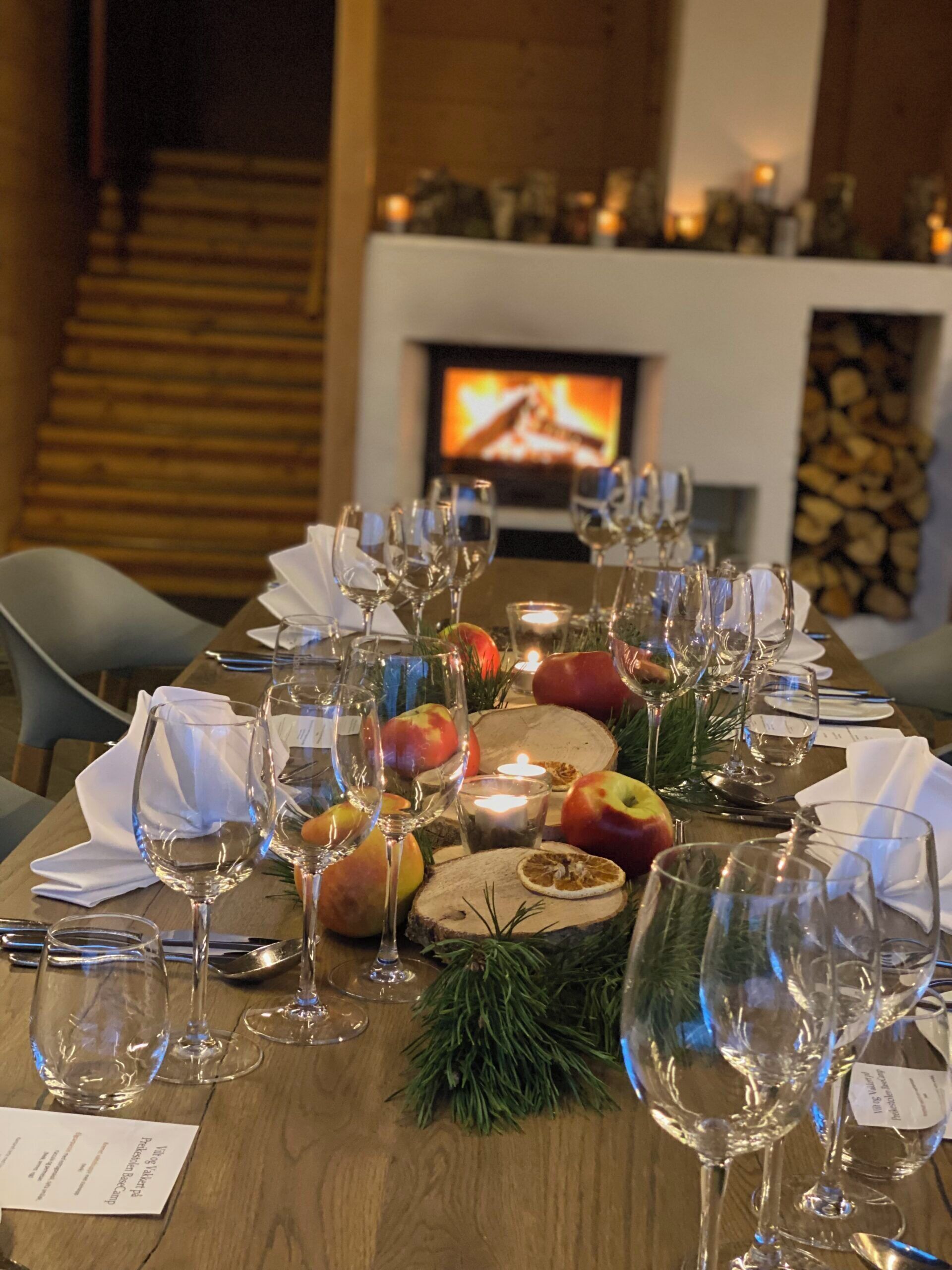 A dining table set with wine glasses, plates, and apples, with a fireplace and television in the background.