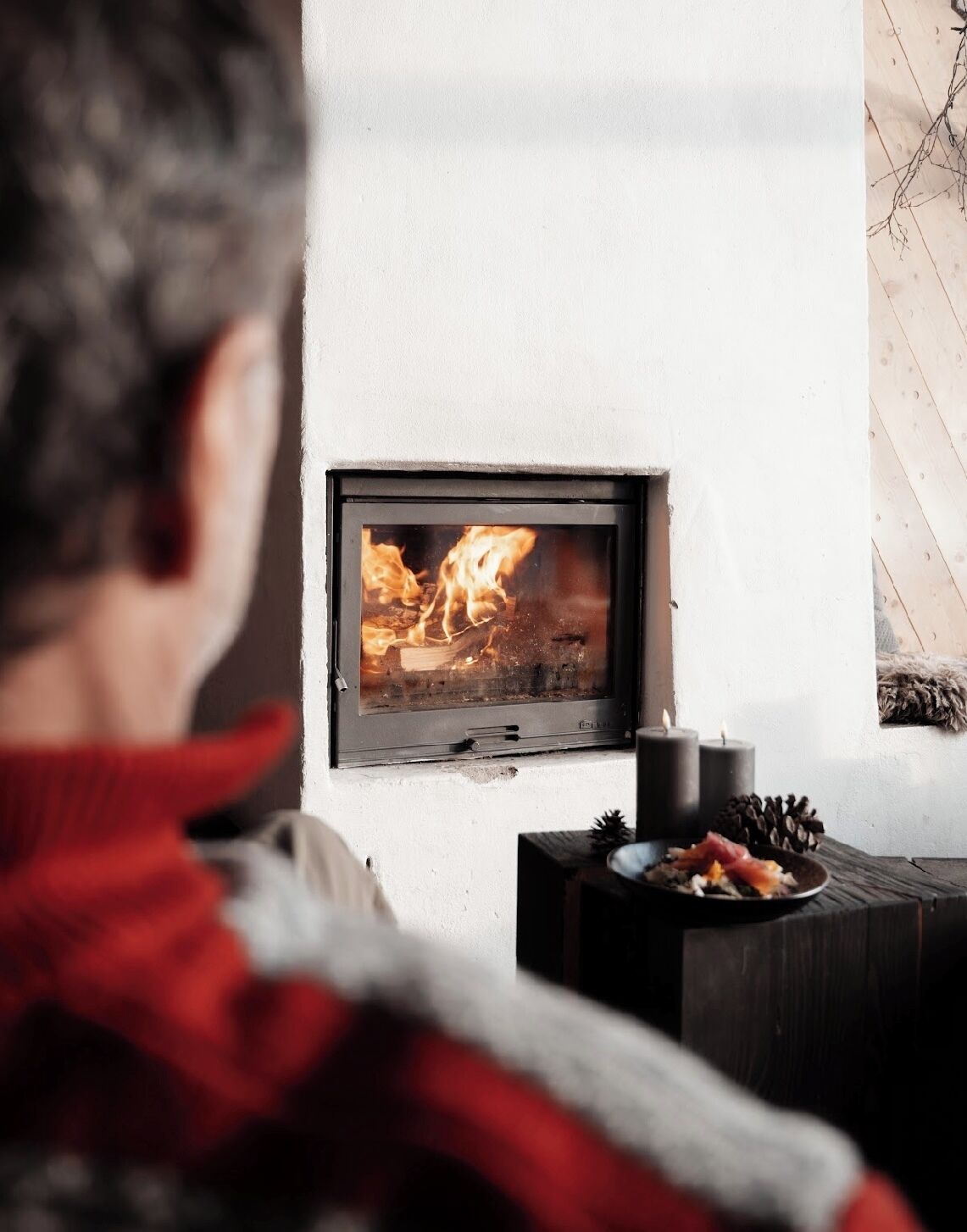 A person sitting in front of a fireplace with a modern design, a small table with candles, and a plate of food.
