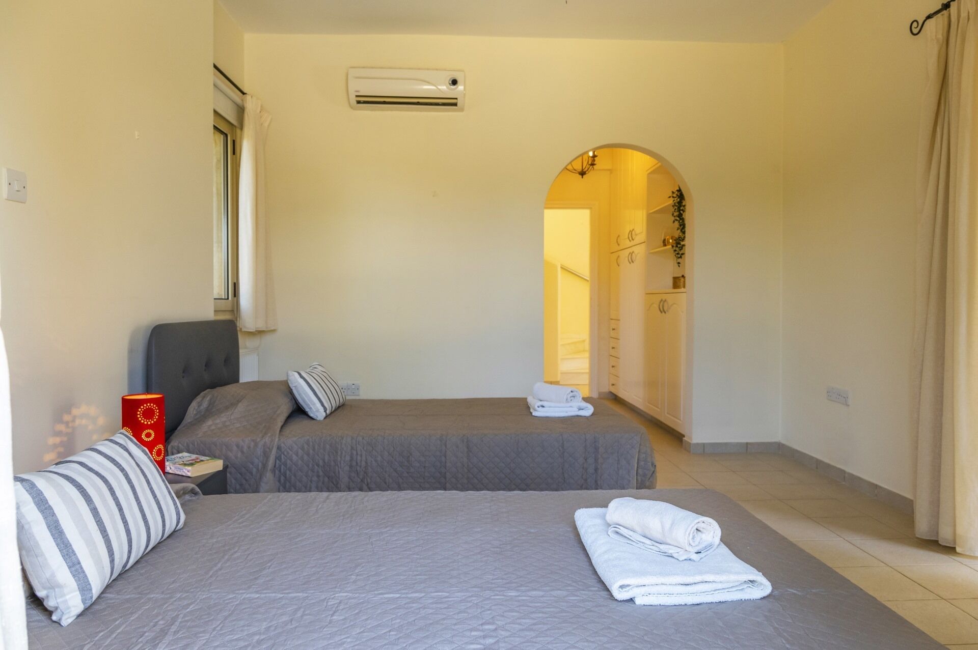 A hotel room with two beds, a window, an air conditioning unit, a red lamp, and a towel on the bed.