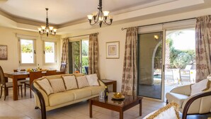 Una sala de estar con sofá, mesa de centro y comedor con sillas y una mesa. Se observa una araña, ventanas amplias y vistas al exterior.