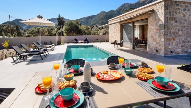 Desayuno junto a la piscina con vistas a montañas, un edificio de piedra y sillones de descanso.