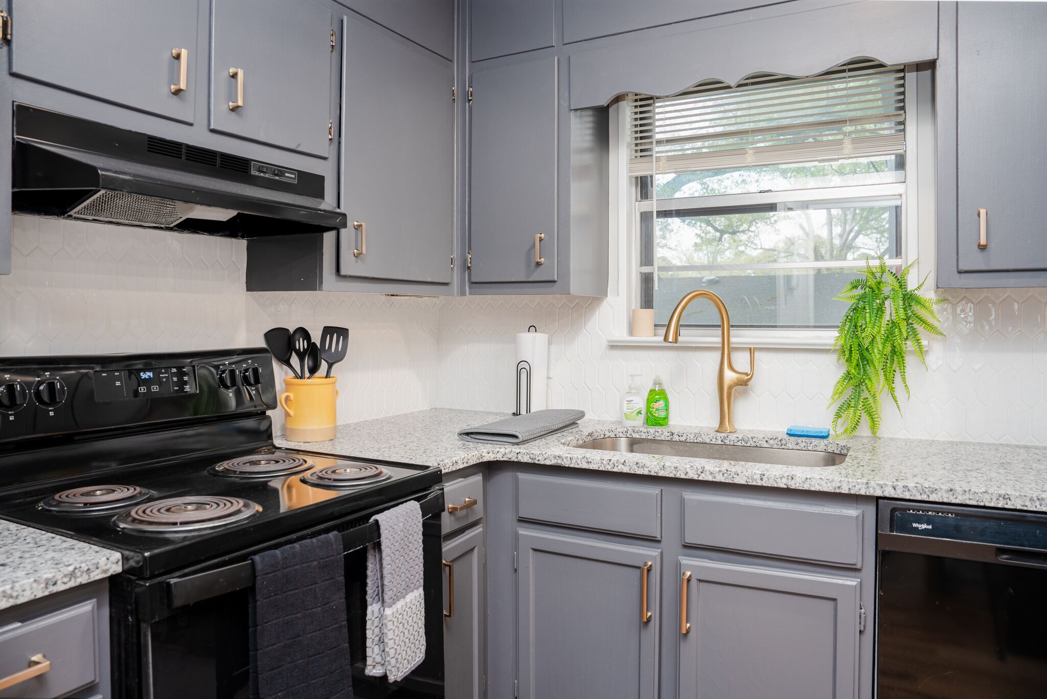 Une cuisine avec des meubles de couleur grise, une cuisinière noire et une fenêtre équipée de stores.