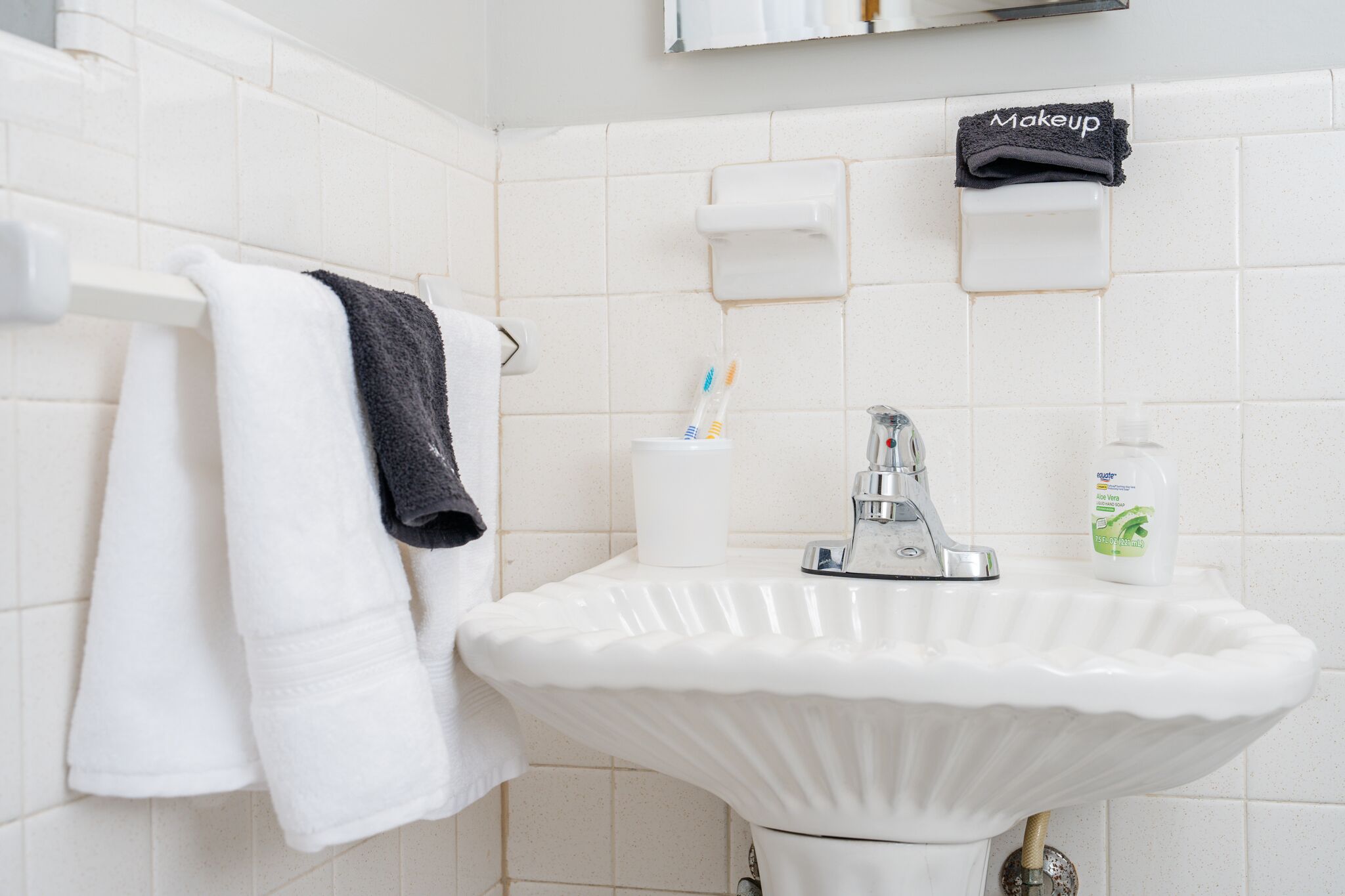 Ett badrum med en vit handfat, en handdukshängare med vita och svarta handdukar, en tvålpump och en handduk för smink som hänger på väggen.