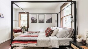A bedroom with a four-poster bed, a bedside table with a lamp, and a window with a view of trees. - Entire Cabin - Modern A-Frame w/Hot Tub (Ellenville)