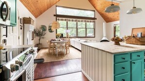 A kitchen with wooden beams, a dining area with a table and chairs, a large window, and a teal-colored island with drawers. - Entire Cabin - Modern A-Frame w/Hot Tub (Ellenville)
