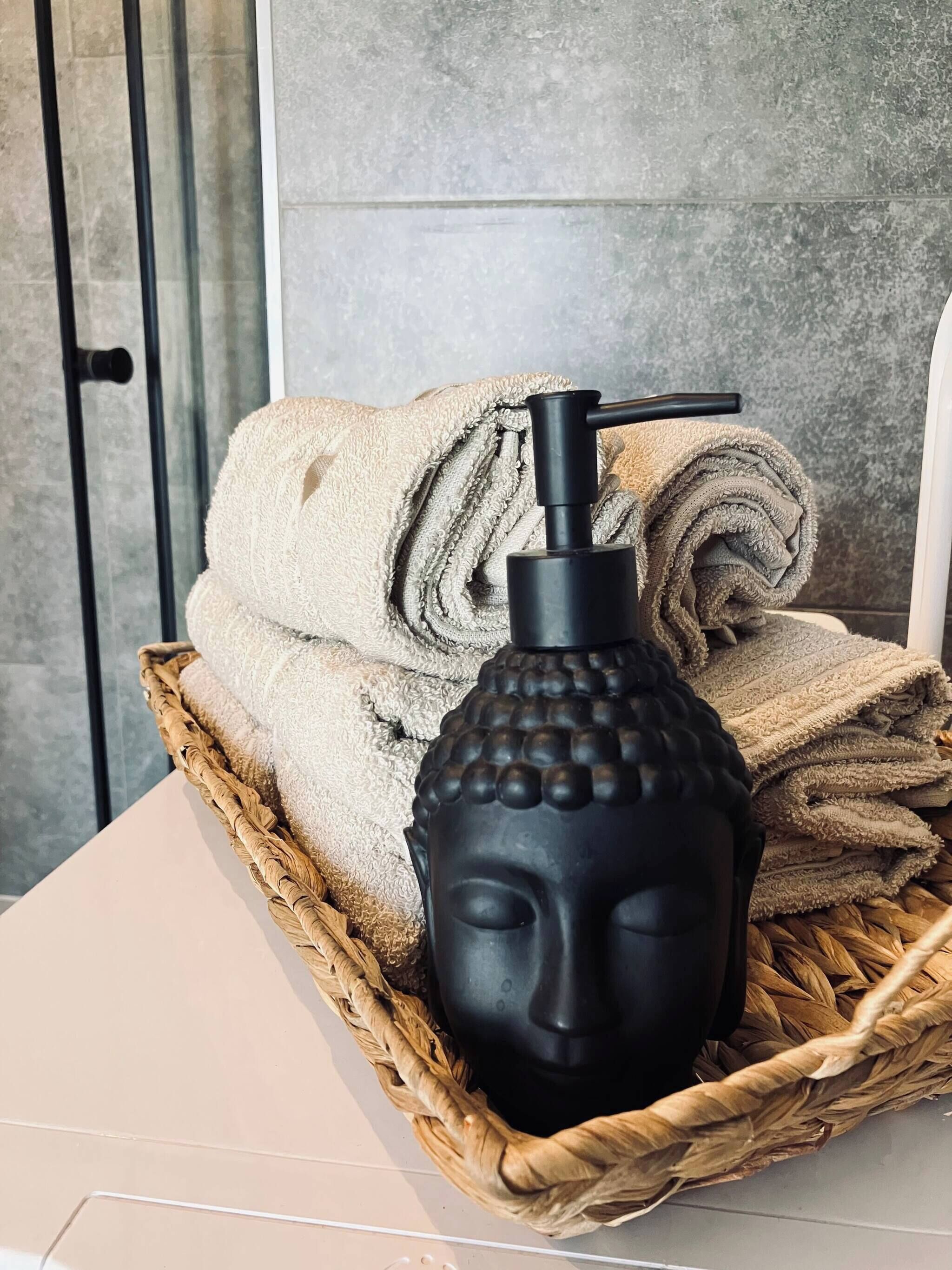 Una cesta tejida con toallas enrolladas y una estatua negra de Buda sobre una mesa blanca.