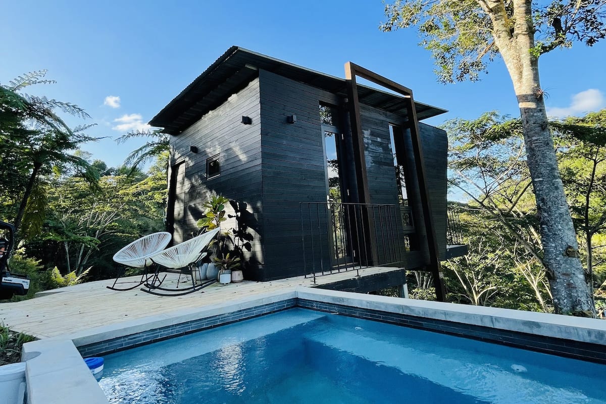 Un área de piscina moderna al aire libre con terraza de madera, dos sillas de descanso blancas y un pequeño edificio con techo oscuro.