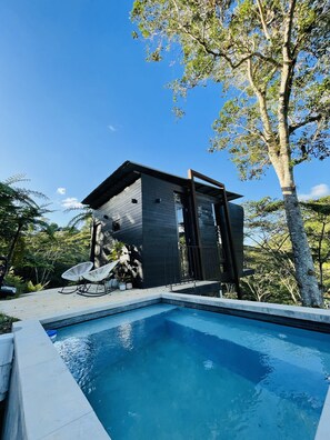 Un área de piscina moderna al aire libre con terraza de madera, dos sillas de descanso blancas y un pequeño edificio con techo oscuro.