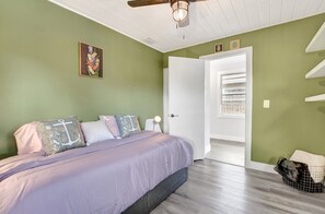Ein Schlafzimmer mit einem Bett, Kissen, einem Nachttisch, einem Deckenventilator, einer Tür und einem an der Wand befestigten Regal.