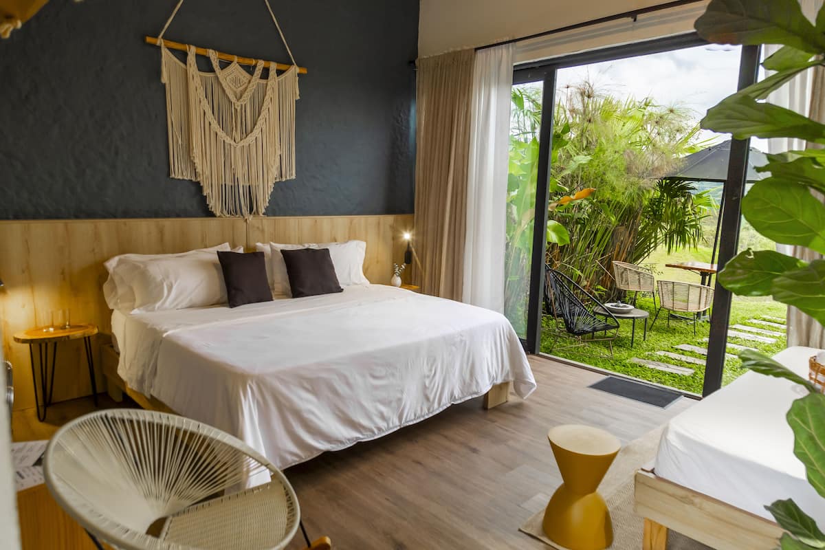 A modern hotel room with a large bed, a macrame wall hanging, and a view of the garden through the sliding glass door.