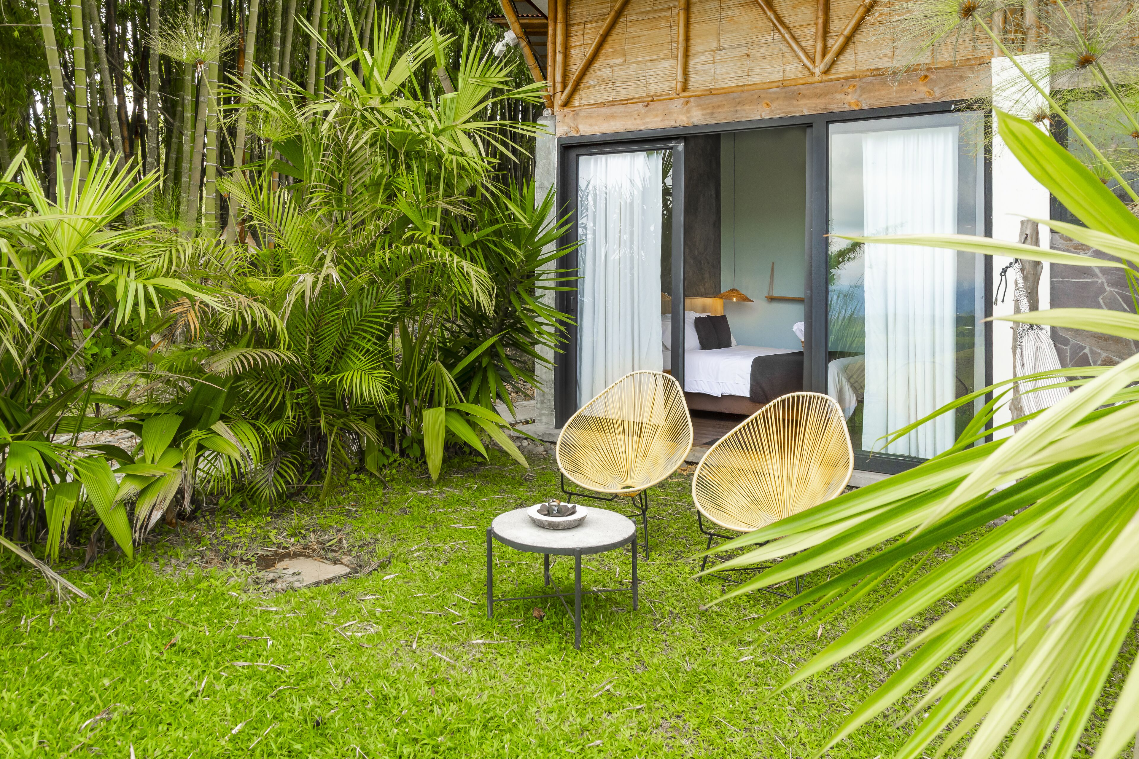A lush garden with bamboo, a small round table with a bowl, and two wicker chairs.