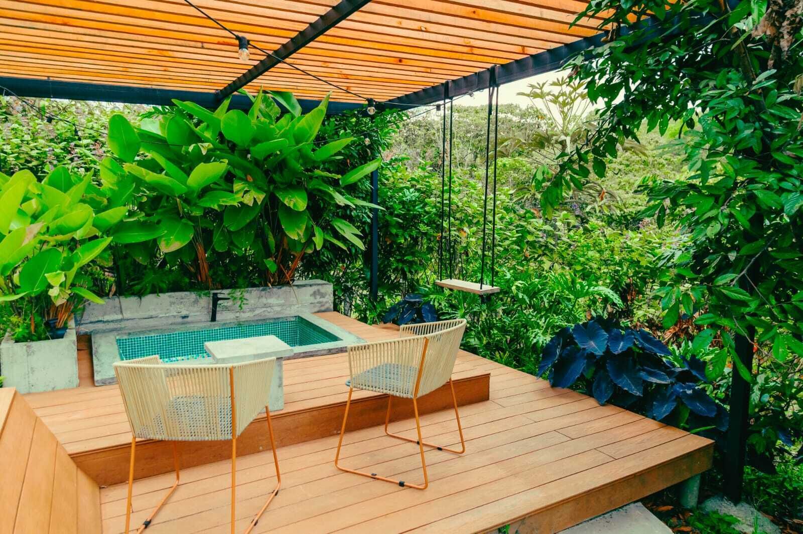 A wooden deck with a pool, two wicker chairs, and lush greenery.