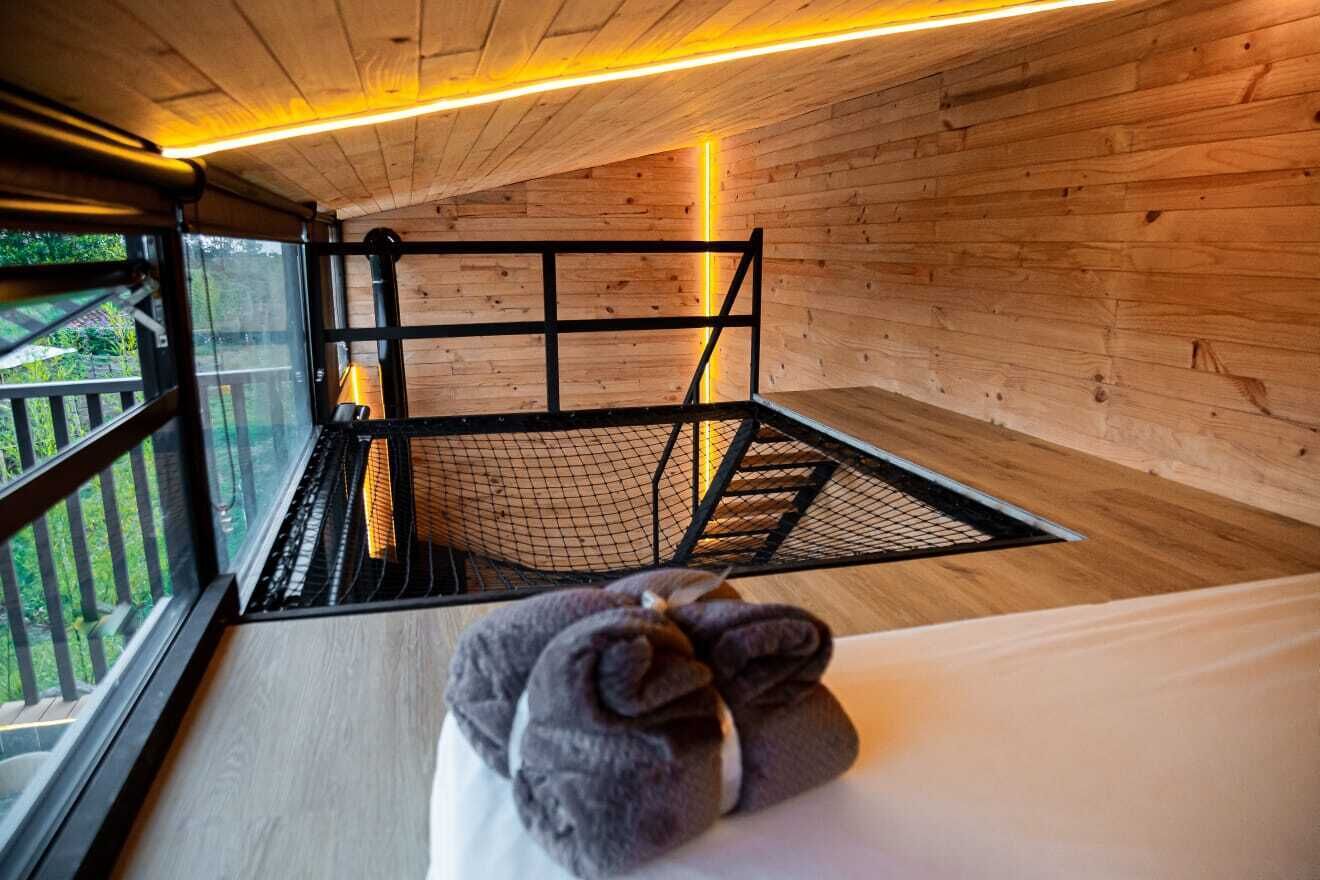 Una habitación de cabaña de madera con literas, ventana con vistas a la vegetación y una lámpara que emite luz cálida.