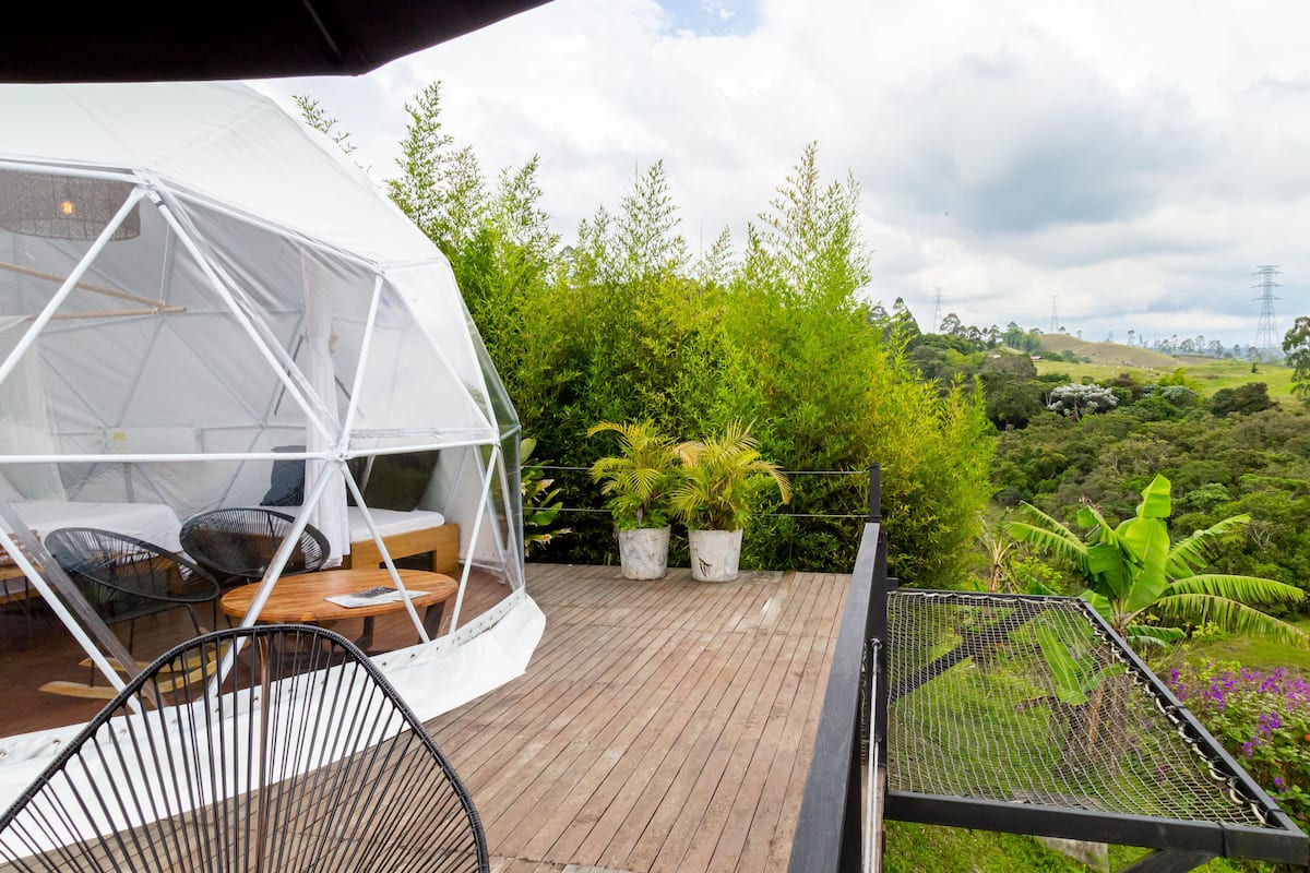 A geodesic dome structure with a wooden table and chairs, surrounded by potted plants and a view of a lush landscape.