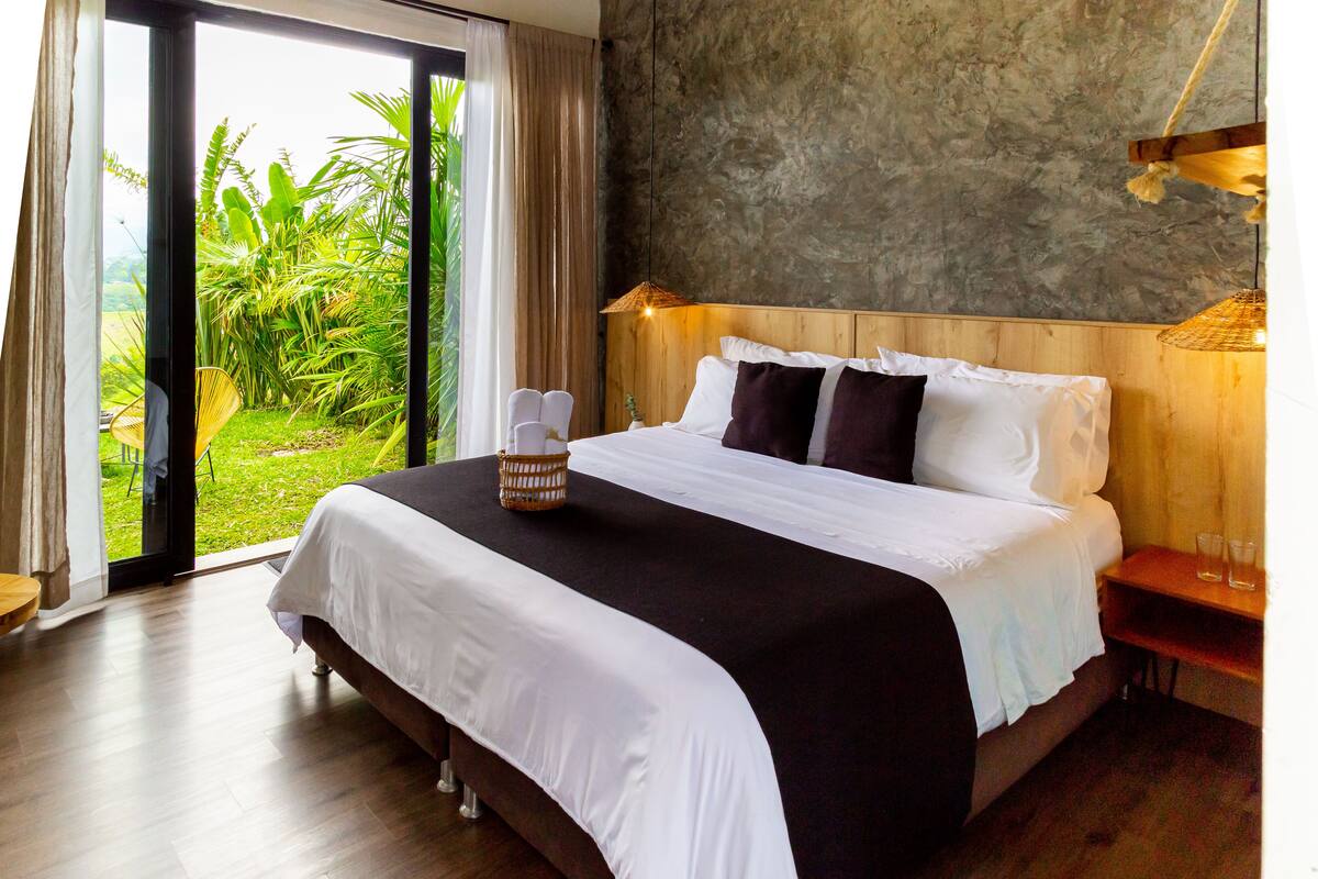 A bedroom with a large bed, a wooden headboard, a view of greenery through a sliding door, and a small bedside table with glasses.