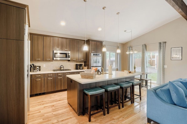 A modern kitchen with wooden cabinets, stainless steel appliances, a central island with bar stools, and a large window providing natural light.