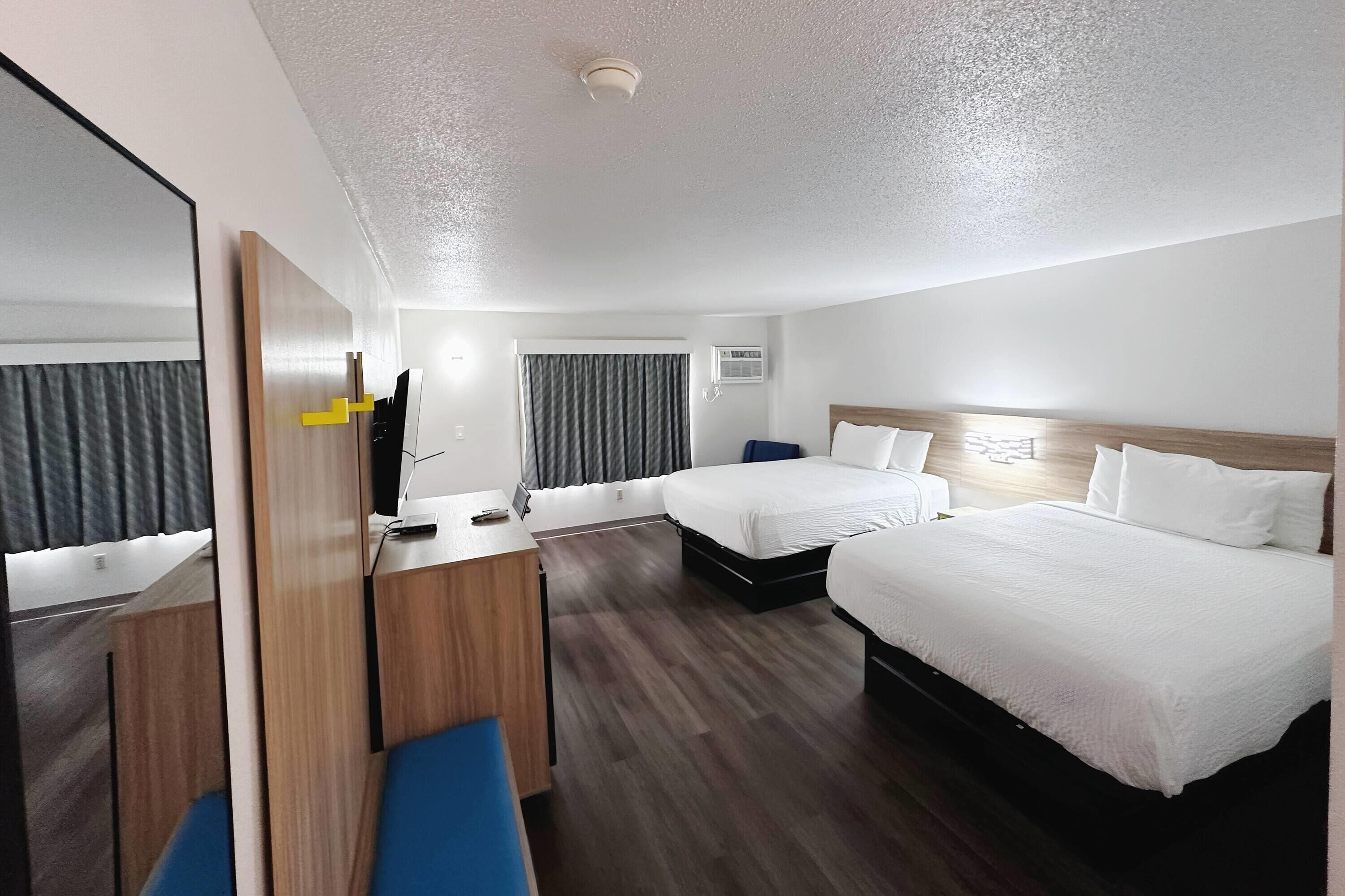 Quarto de hotel com duas camas, uma escrivaninha, uma cadeira, um espelho e uma luminária no teto.