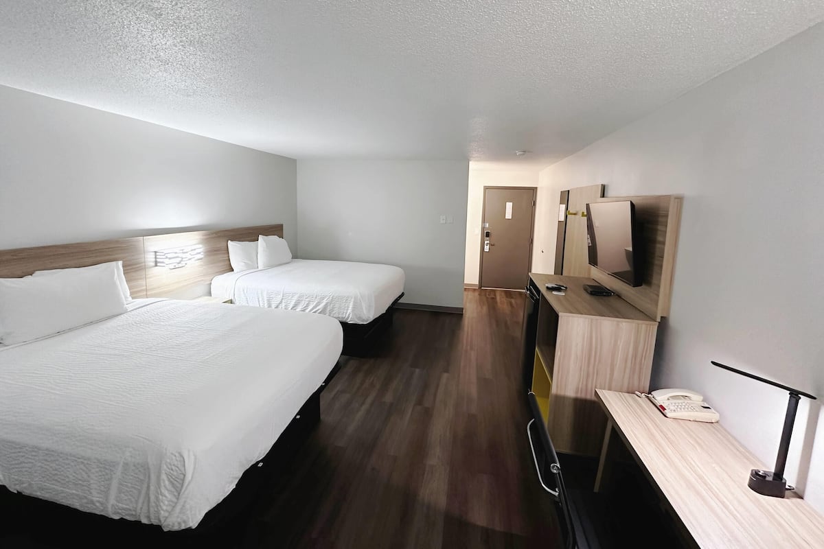 Quarto de hotel com duas camas, uma mesa com telefone e luminária, e uma televisão na parede.