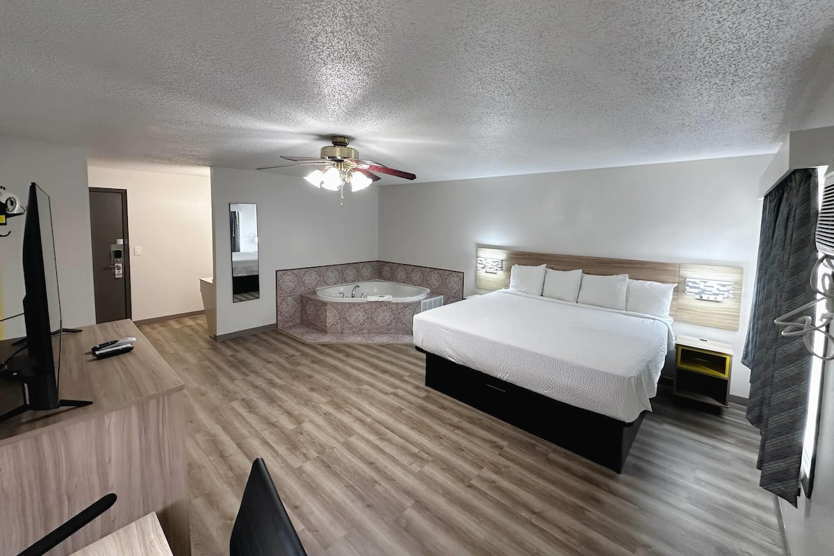 Quarto de hotel com cama, escrivaninha, televisão, ventoinha no teto, banheiro com banheira e janela com cortinas.