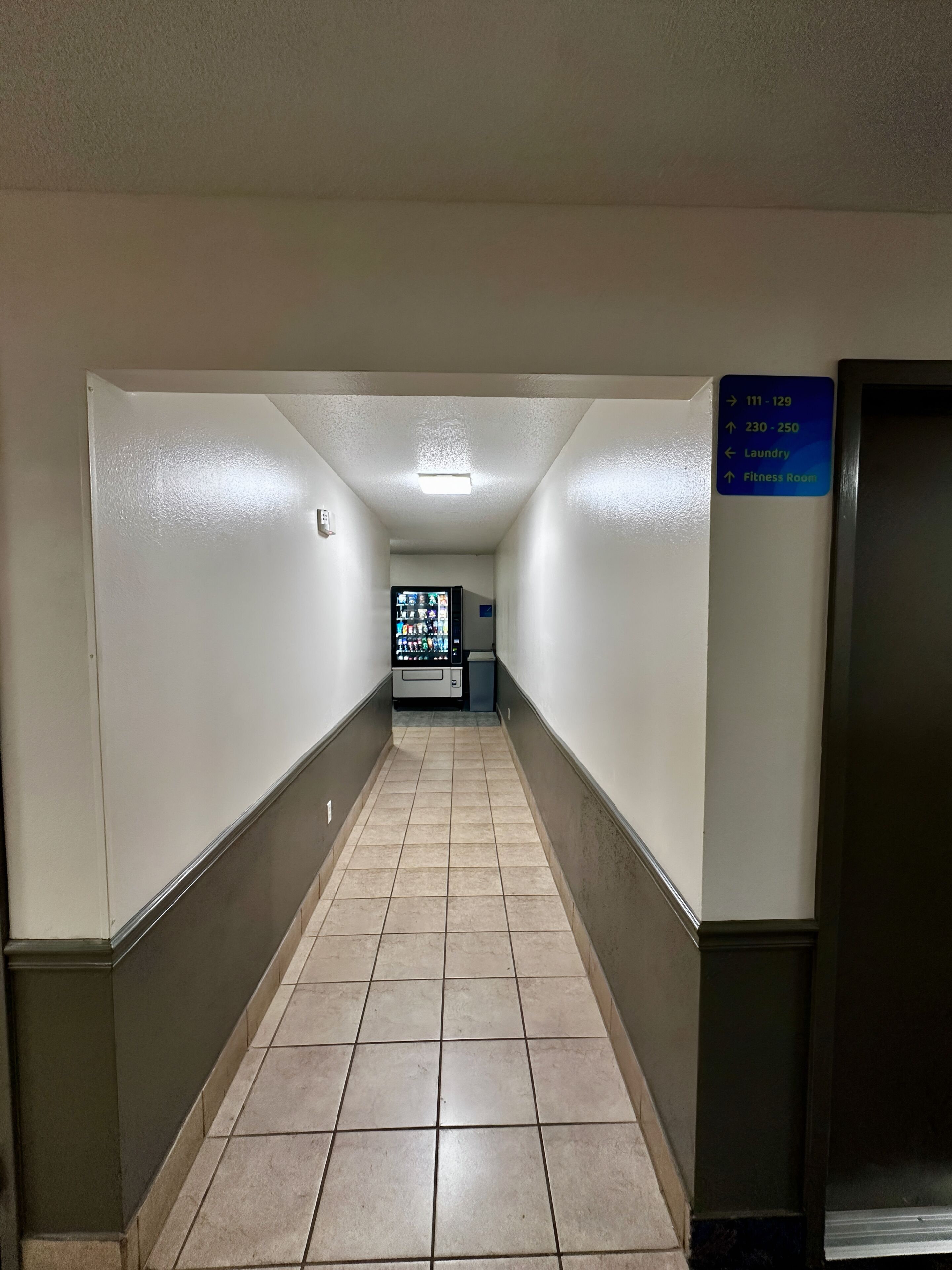 A hotel corridor with a vending machine, a sign indicating room numbers and amenities, and a door on the right.
