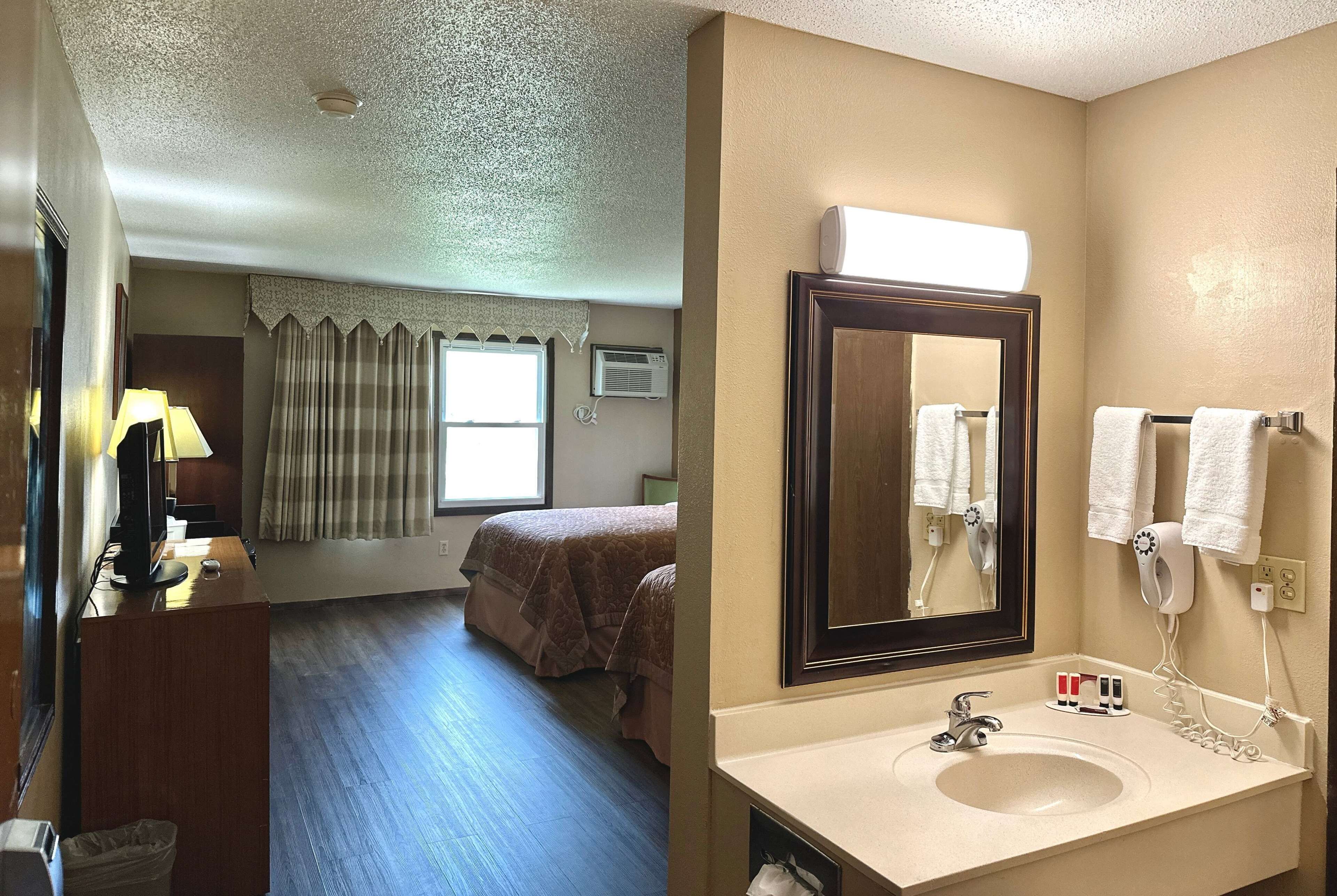 Una habitación de hotel con cama, escritorio con lámpara, baño con lavamanos, espejo y toallero.