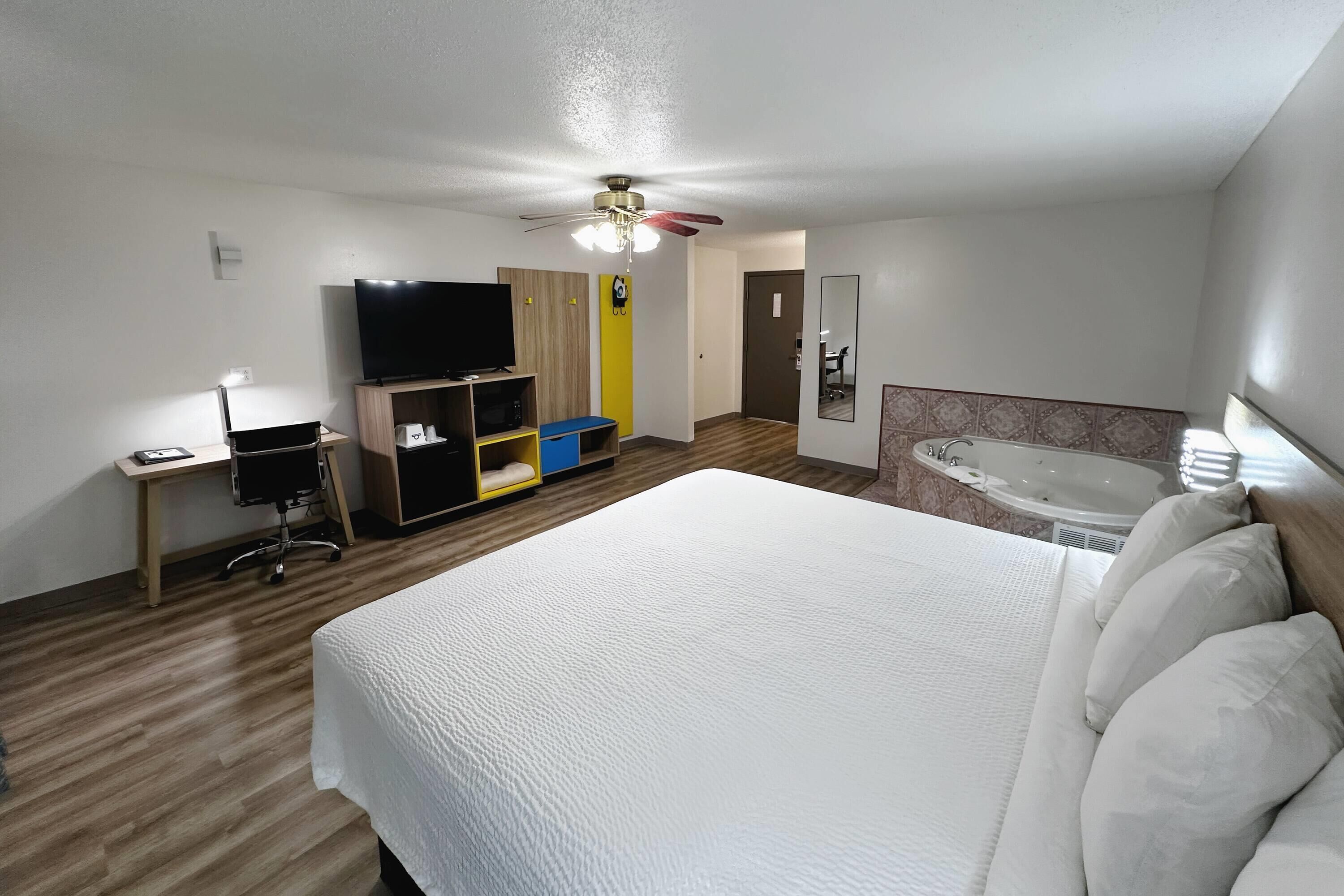 Quarto de hotel com uma cama grande, uma escrivaninha com um computador, uma televisão, um ventilador de teto e uma banheira.