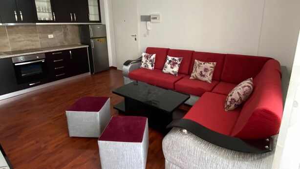 A modern living room with a red sofa, two grey ottomans, a black coffee table, and a kitchen area with dark cabinets and a white door. - Albania Dream Holidays Accommodations (Sarandë)
