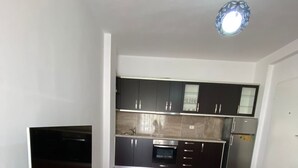 A modern living room with a flat-screen TV, a sofa, and two square ottomans with red cushions. The room has wooden flooring and a kitchen in the background with dark cabinets and stainless steel appliances. - Albania Dream Holidays Accommodations (Sarandë)
