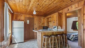 A wooden cabin interior with a kitchen area, a refrigerator, a bar stool seating, and a bed in the background. - Cabin #10 2BR 1BA Timberlane Resort (Park Rapids)