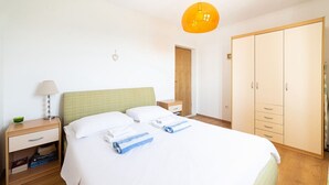 Ein ordentlich gemachtes Bett mit weißen Leinen und blauen Handtüchern, ein Nachttisch mit Lampe, ein Holzbeistelltisch, ein Kleiderschrank und eine Tür zu einem anderen Raum.