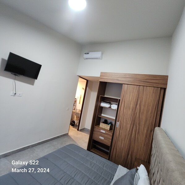 A bedroom with a bed, a TV mounted on the wall, a wardrobe, and an air conditioning unit.
