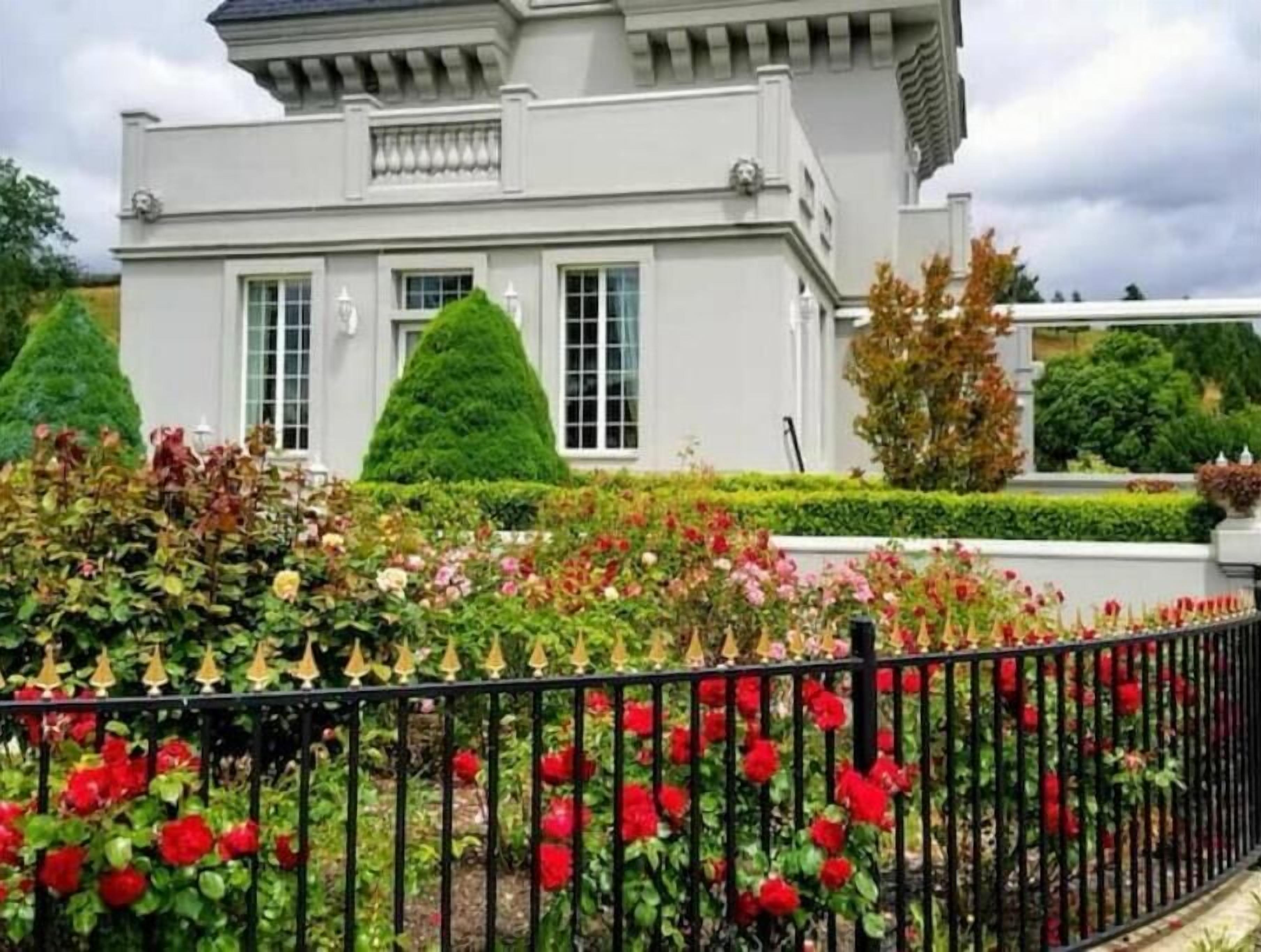 Um edifício com fachada simétrica, grandes janelas e um jardim bem cuidado com flores vermelhas e sebes aparadas.