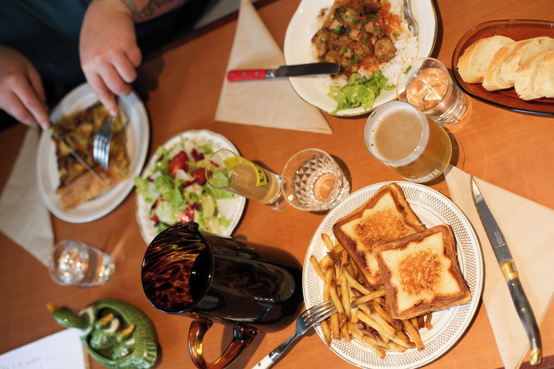 A dining table with various dishes, including a plate of French fries, a sandwich, and a salad. There are also drinks and utensils on the table.