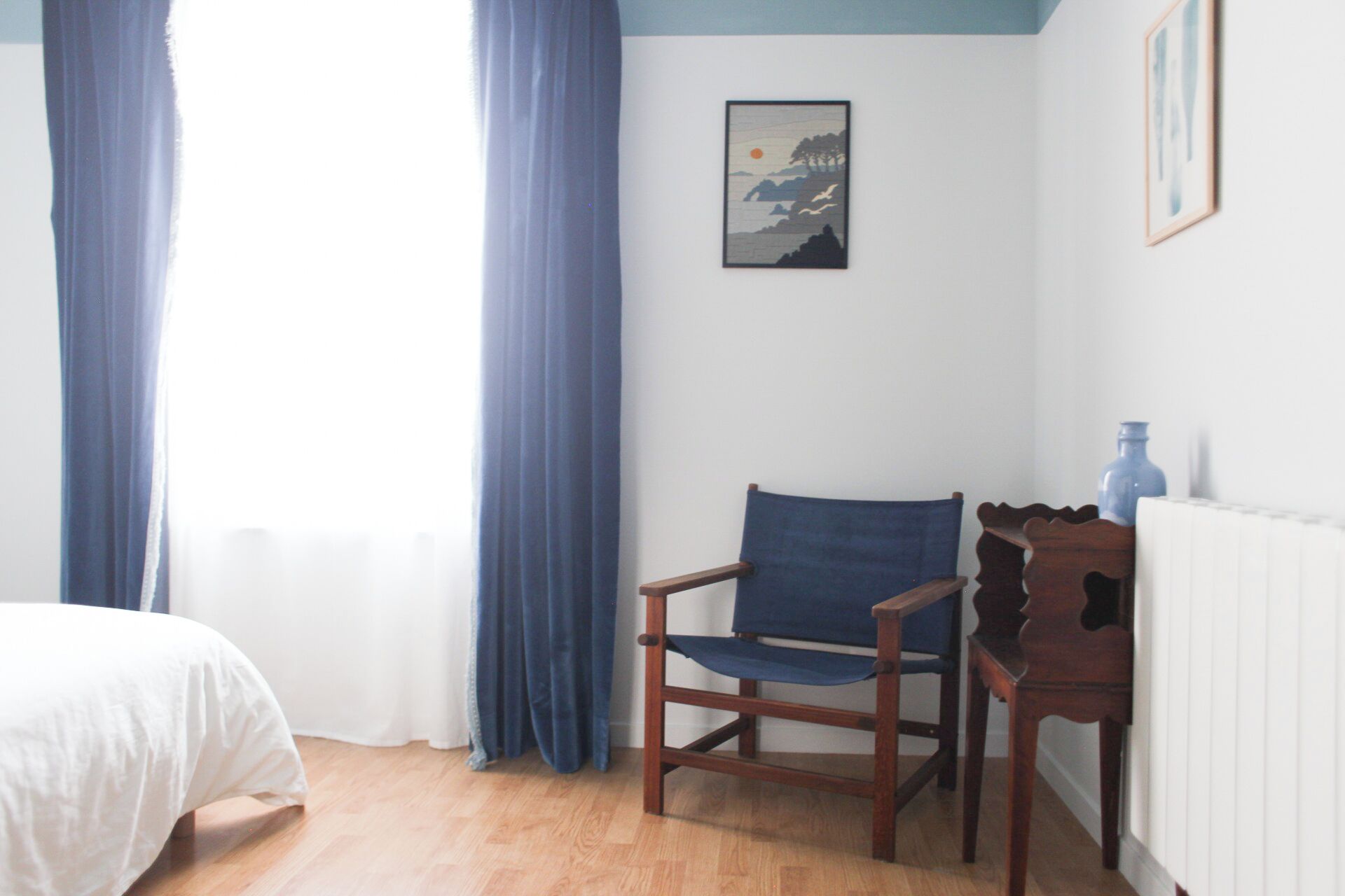 A room with a wooden chair, a bed, a radiator, and a framed picture on the wall.