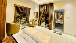 Hotelkamer met een bed, twee fauteuils, een bijzettafeltje, een plafondventilator en een zichtbare badkamer door een glazen deur.