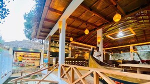 Een restaurant met een houten terras, binnenzitplaatsen en een rieten dakstructuur op de achtergrond.