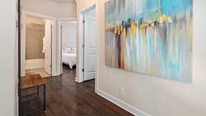 A hallway with a wooden floor, a large abstract painting on the wall, and a glimpse of a bedroom with a bed and a bathroom with a shower and bathtub. - Chic 3-Bed Condo, 2-Car Garage close to Pearl & St. Mary's Strip in Tobin Hills! (San Antonio)