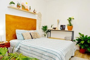 Une chambre à coucher avec une tête de lit en bois, une couvre-lit à rayures, plusieurs coussins, une table de chevet, une commode, une étagère avec des plantes et un tableau encadré.
