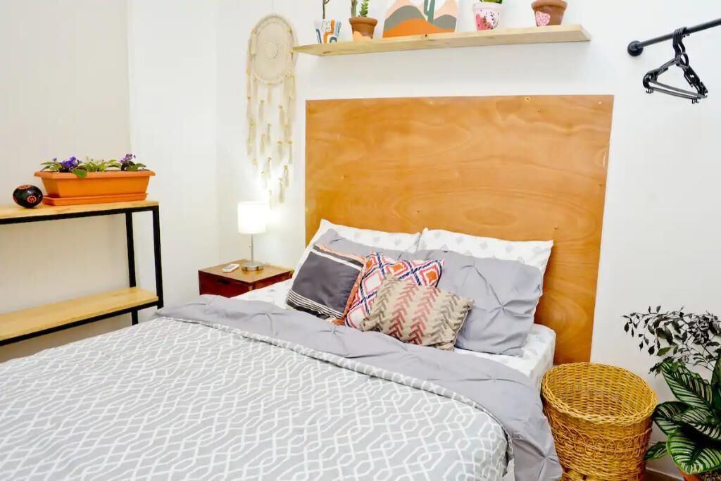 A neatly arranged bedroom with a wooden headboard, a bedside table with a lamp, and a shelf with decorative items.