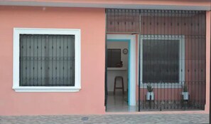 Un bâtiment à la façade rose, une fenêtre ornée d’une grille métallique et une porte donnant sur un espace intérieur.