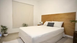 Una cama bien tendida con cabecera de madera, dos mesitas de noche con lámparas y dos macetas con plantas.
