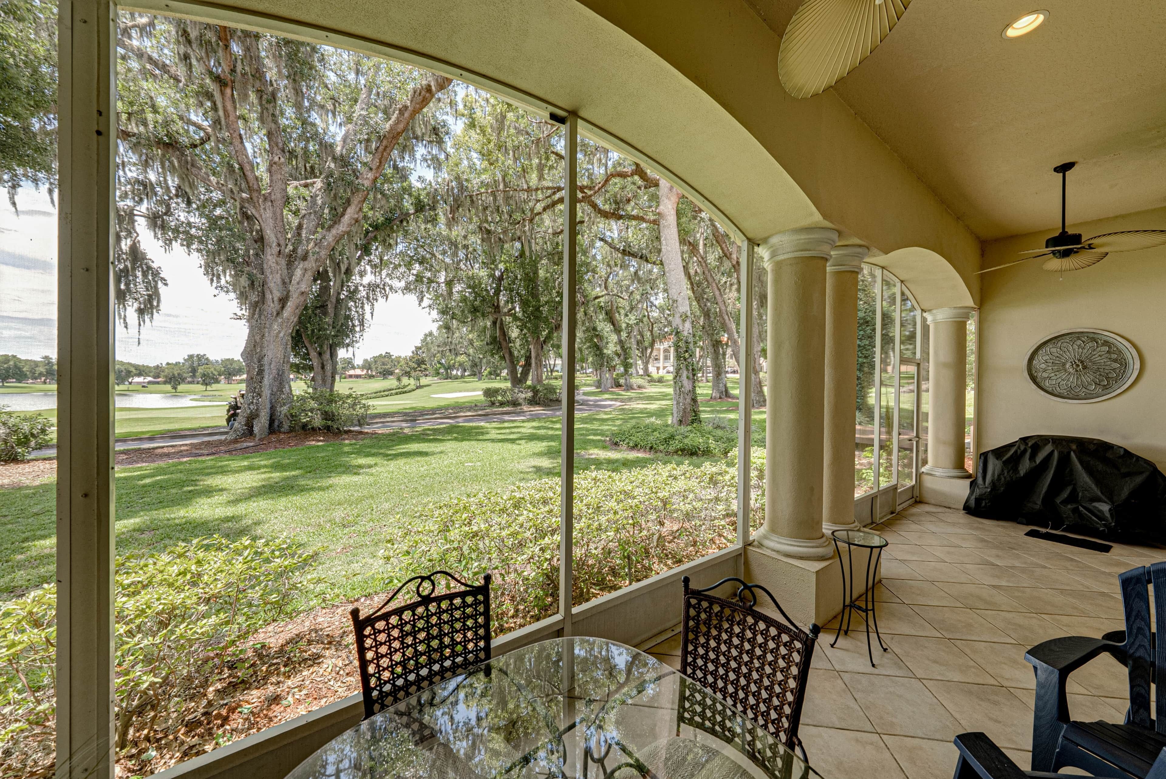A covered patio with a glass table and chairs, overlooking a well-manicured lawn and trees.