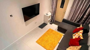 A modern living room with a flat-screen TV mounted on the wall, a dark-colored sofa with red and beige cushions, and a yellow rug on the floor. - 1bedhouse- London-sleeps 4- Close to Attractions (London)